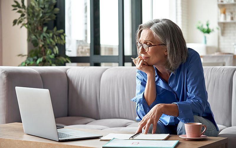 Elderly woman sitting on her sofa looking at paperwork and her laptop at her account