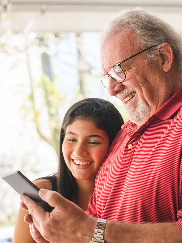 elderly man laughing while using a smartphone with a young woman relative looking at the phone with him laughing