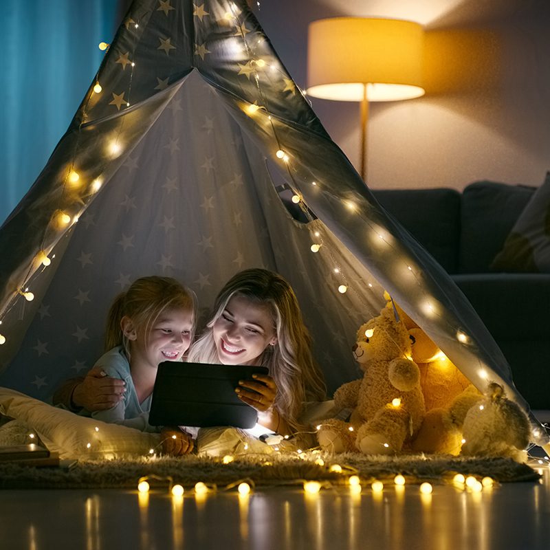 A mother and a daughter watching something on their tablet in a makeshift tent streamed with lights in the living room.